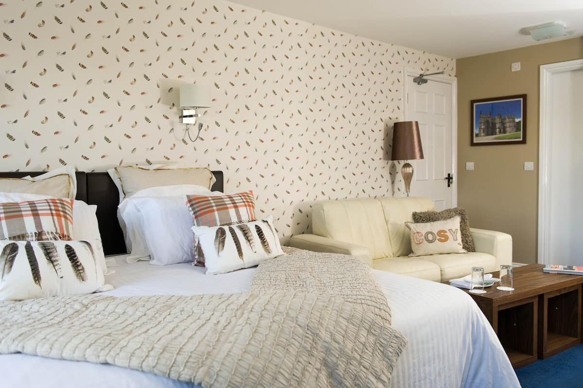 A hotel room with a large bed, a sofa, and a small table. There is a wall-mounted lamp and a picture on the wall.