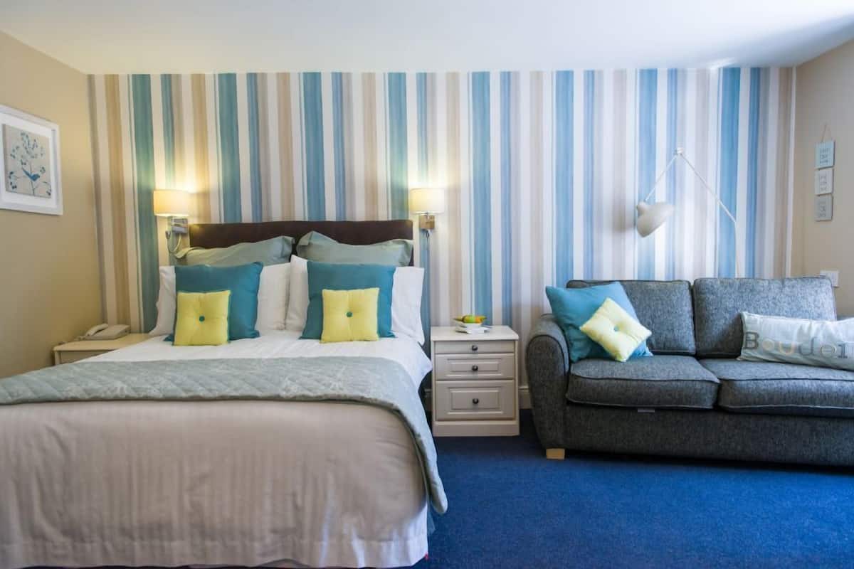 A hotel room with a large bed, a smaller bed, a sofa, striped wallpaper, and a nightstand with a lamp.