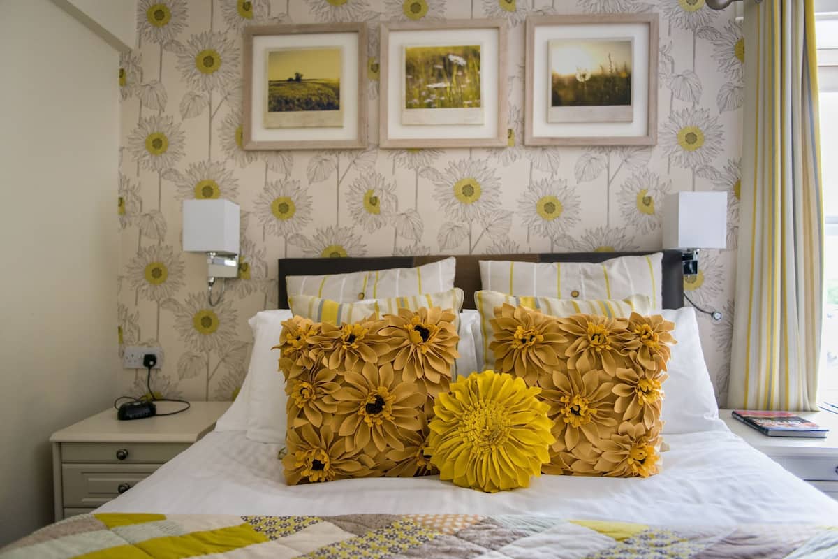 A neatly made bed with floral-patterned pillows and a quilt, a bedside table with a lamp, and framed pictures on the wall.