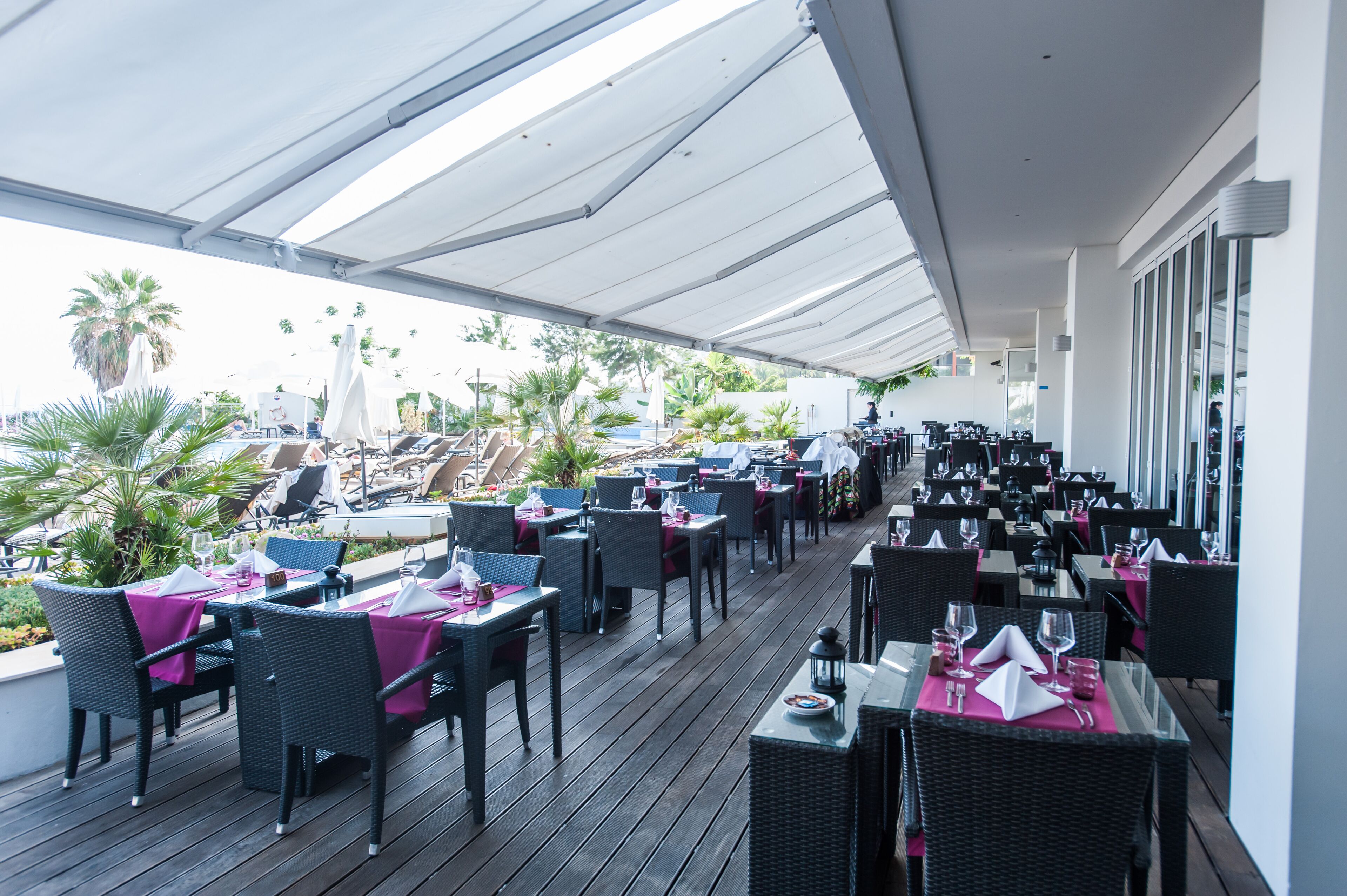 An outdoor dining area with tables and chairs, some with umbrellas, and a view of a pool and palm trees.