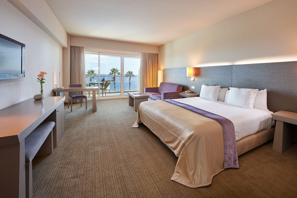 A hotel room with a large bed, a desk, a chair, a table, and a view of the ocean.