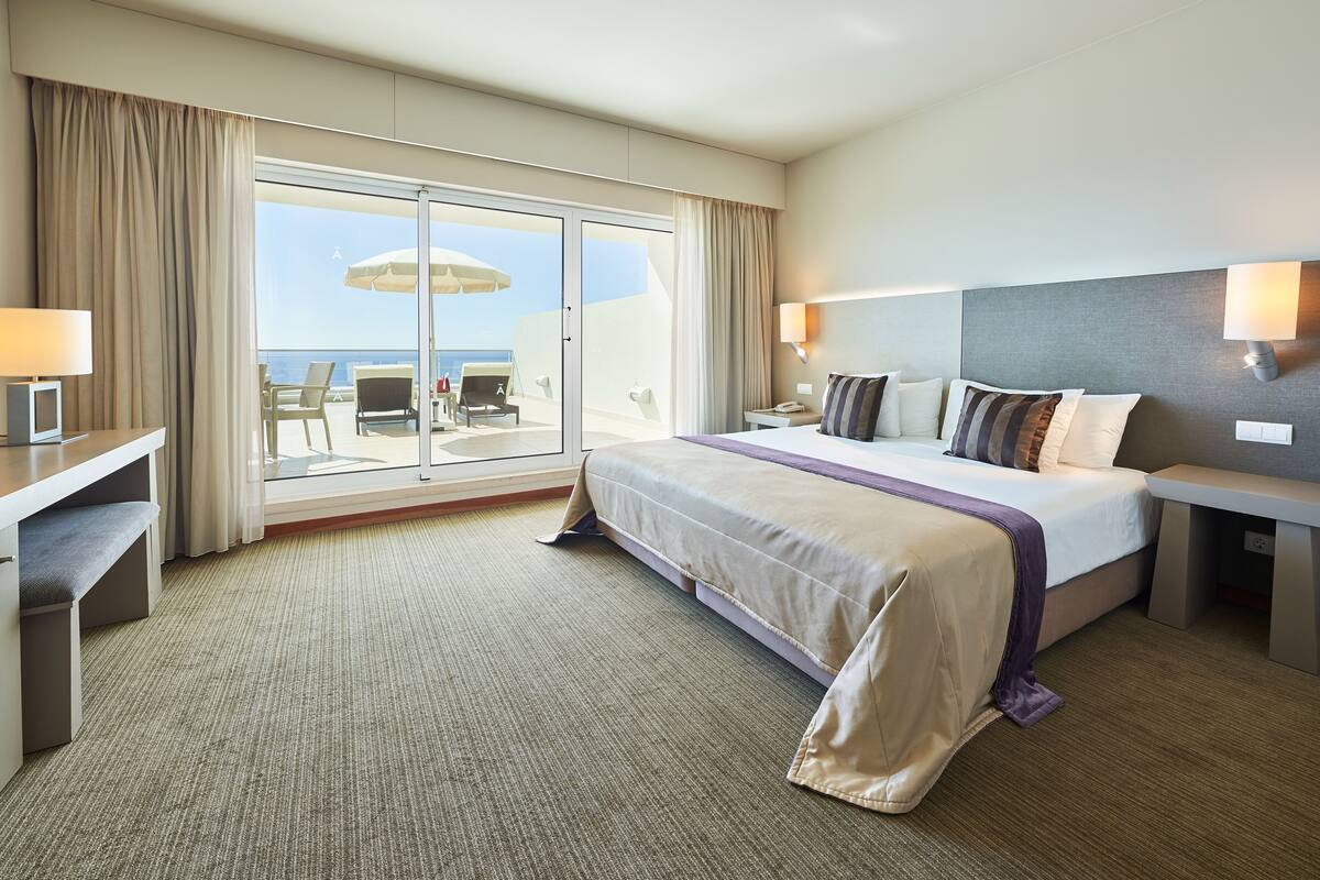 A hotel room with a large bed, a desk, a chair, and a view of the ocean through a sliding glass door.