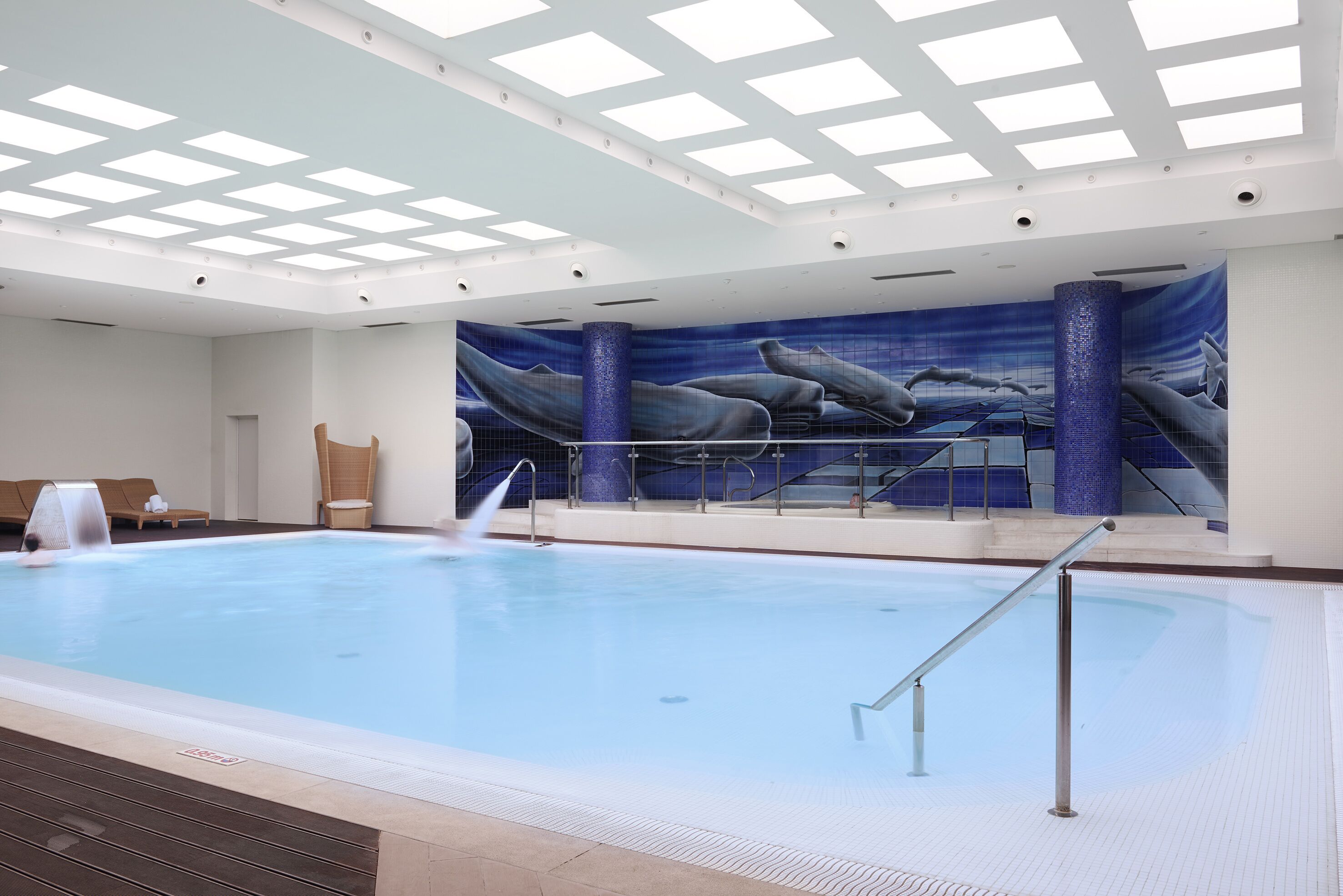 An indoor swimming pool with a mural of whales on the wall, a sunroof, and a lounge area with chairs.
