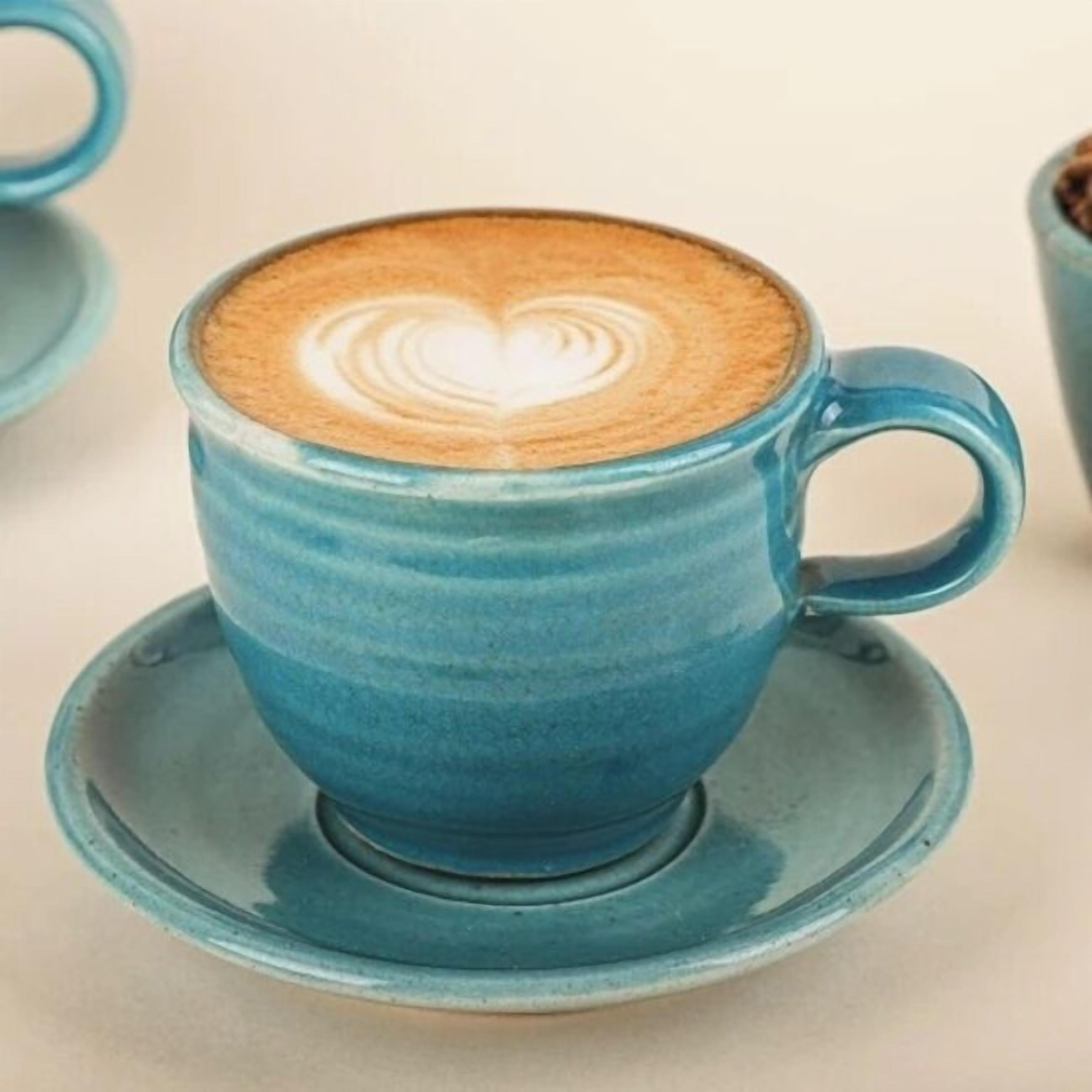 A blue ceramic cup with a saucer, containing a latte with a heart-shaped latte art on the foam.