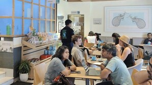 Interior moderno de una cafetería con vigas de madera en el techo, mesas y sillas redondas. Se ve una vitrina con productos de panadería.