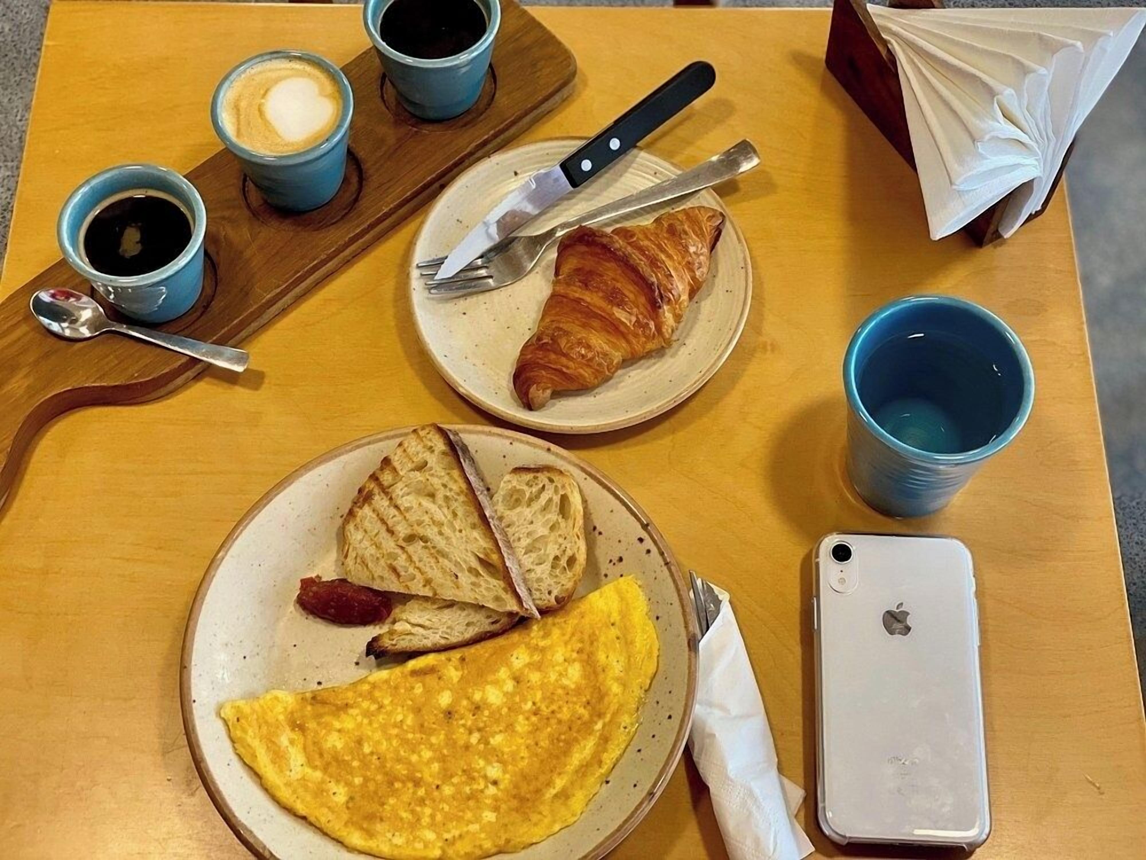 A breakfast setting with croissants, toast, an omelette, coffee, and a smartphone on a wooden table.