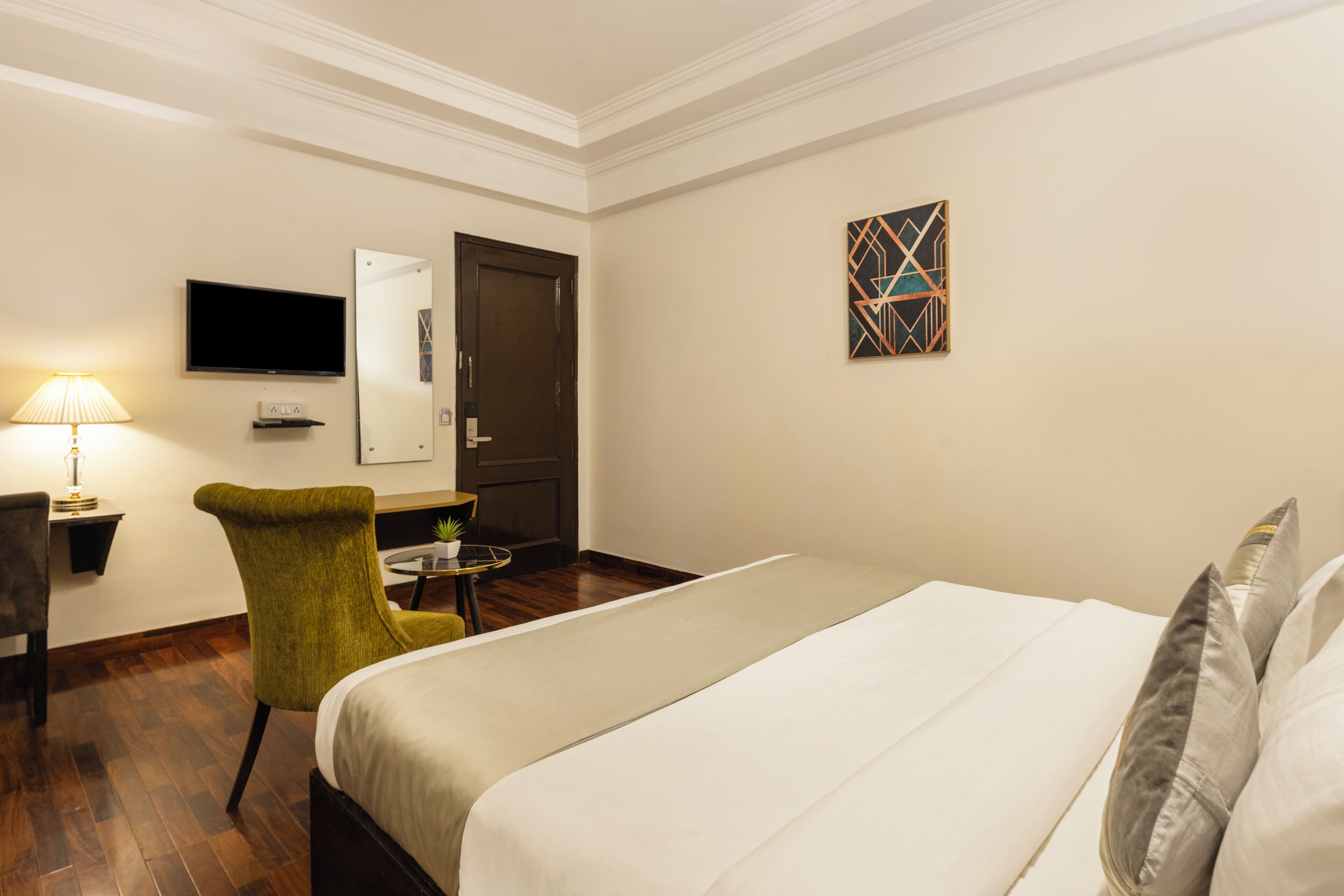 A hotel room with a bed, a green armchair, a small table with a lamp, a TV, and a door.