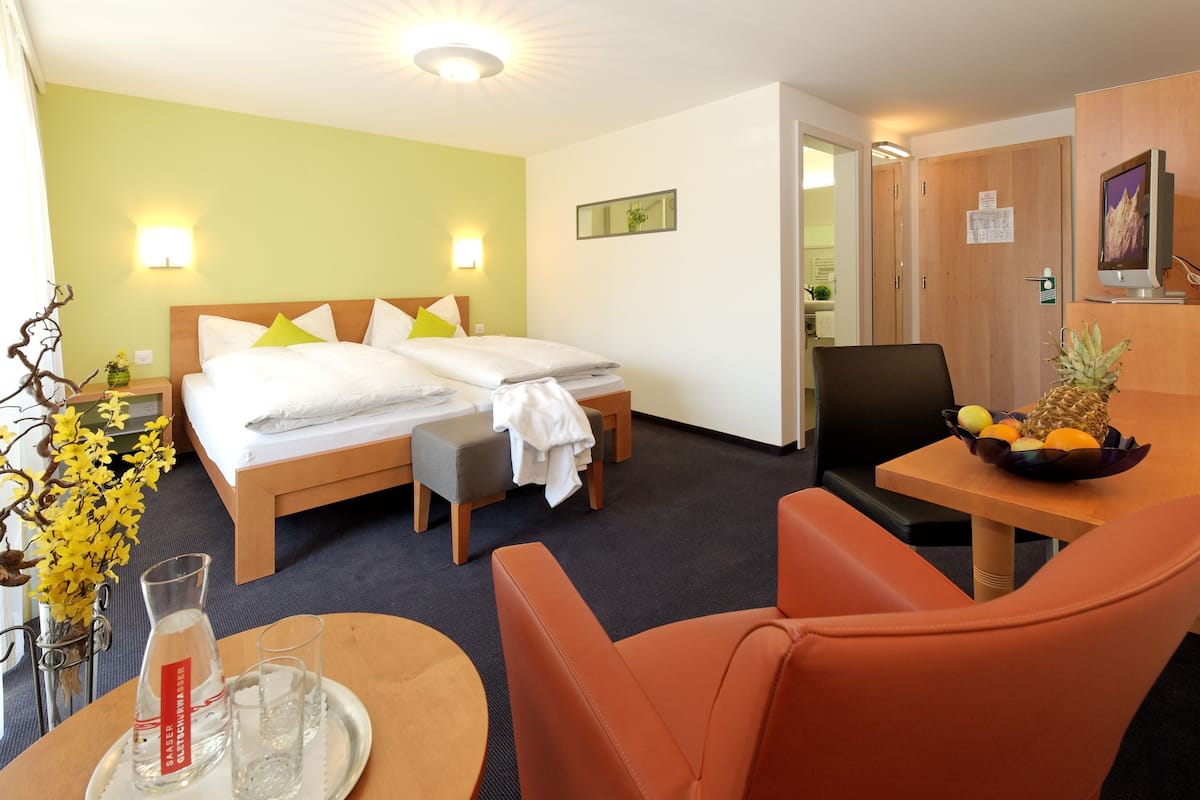 A hotel room with a bed, a desk with a fruit bowl, a chair, and a table with a water bottle and glasses.