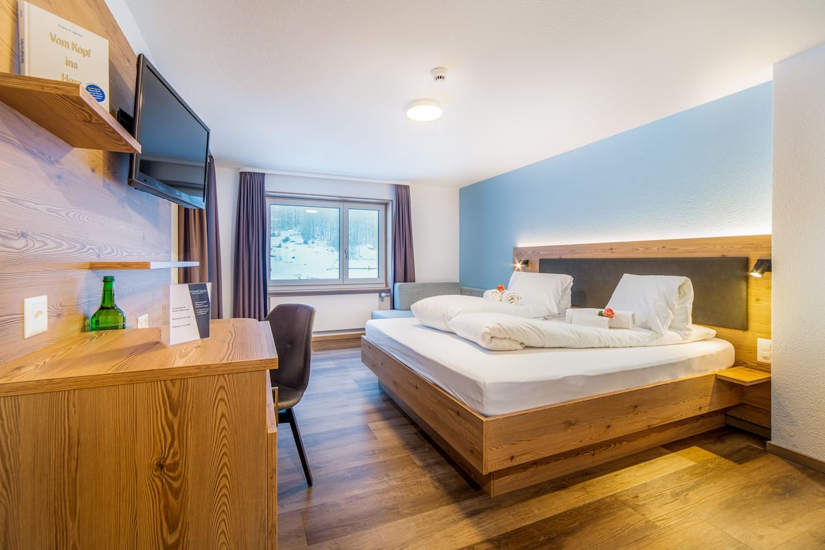 A hotel room with a wooden desk, a bed with white linens, a television mounted on the wall, and a window with a view of a snowy landscape.