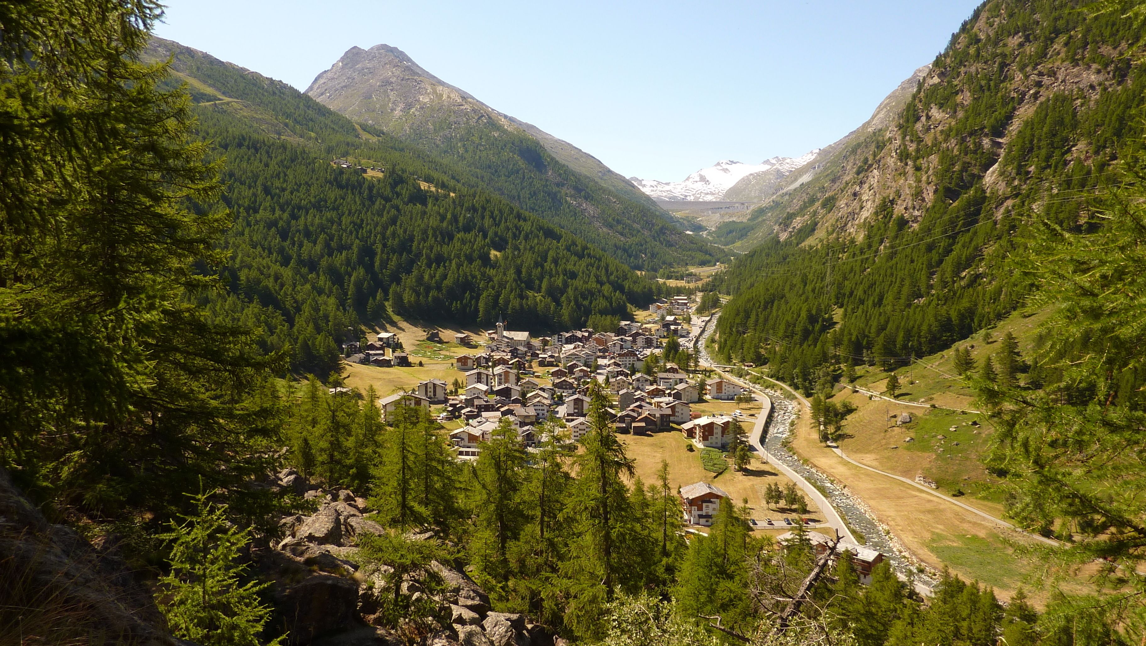 Un pueblo montañoso con casas y un río, rodeado de densos bosques y cumbres nevadas.