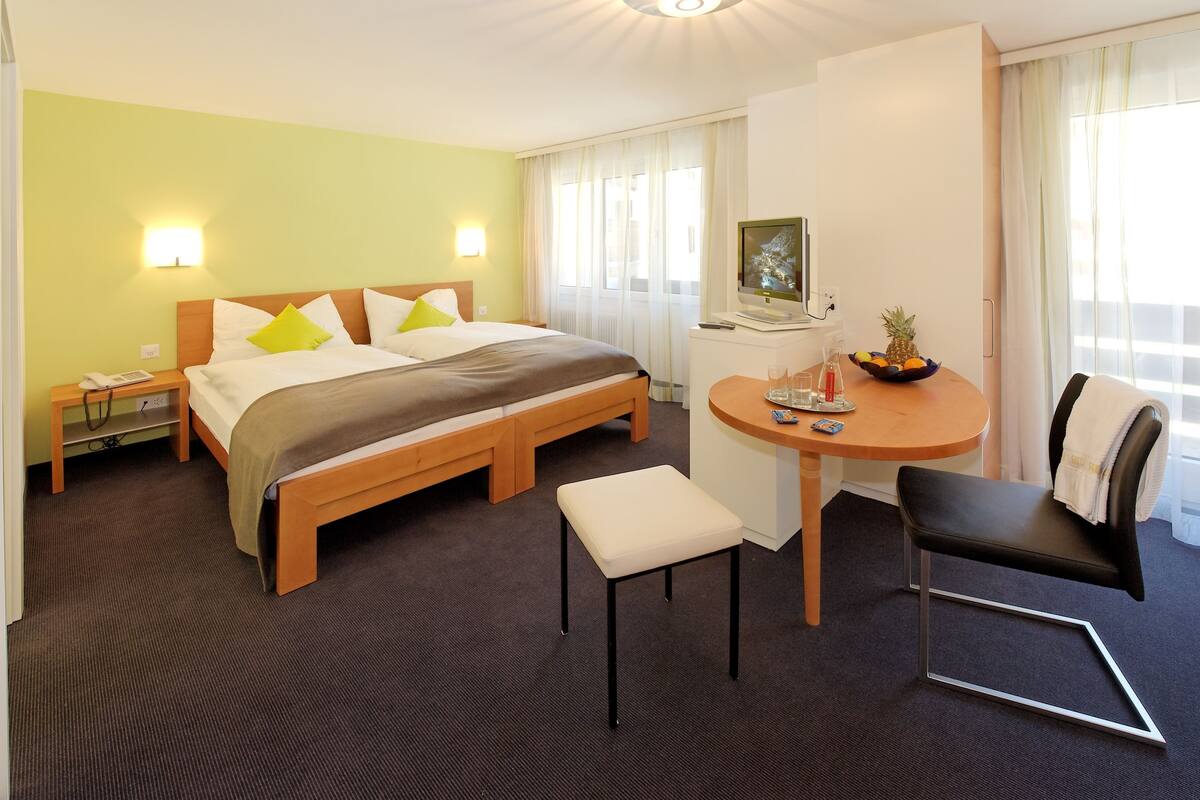 A hotel room with a bed, bedside table, chair, desk, and a small table with a fruit bowl.