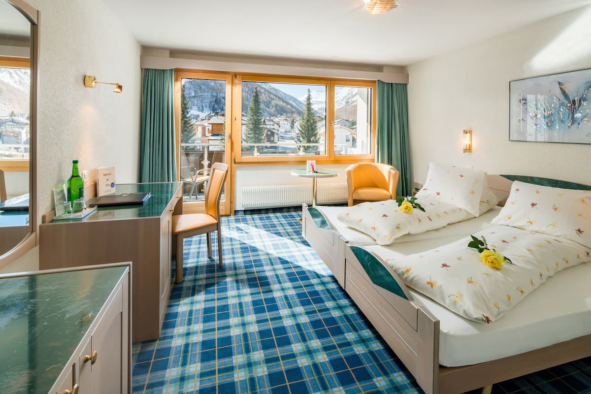 A hotel room with a bed, a desk, a chair, a small table, and a view of mountains through large windows.