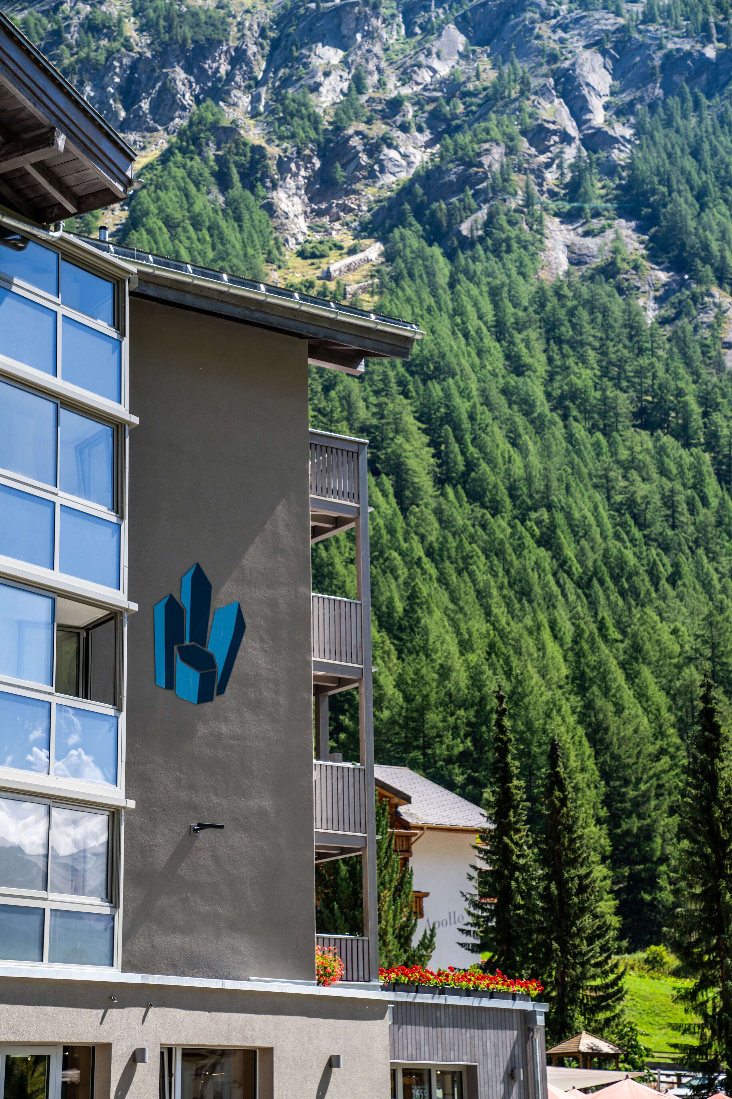 A hotel with a modern design, featuring a prominent logo on the facade, balconies, and a view of a forested mountain range.