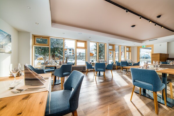 Un restaurante moderno con mesas de madera y sillas azules, amplios ventanales que ofrecen vistas al exterior y un interior limpio y luminoso.