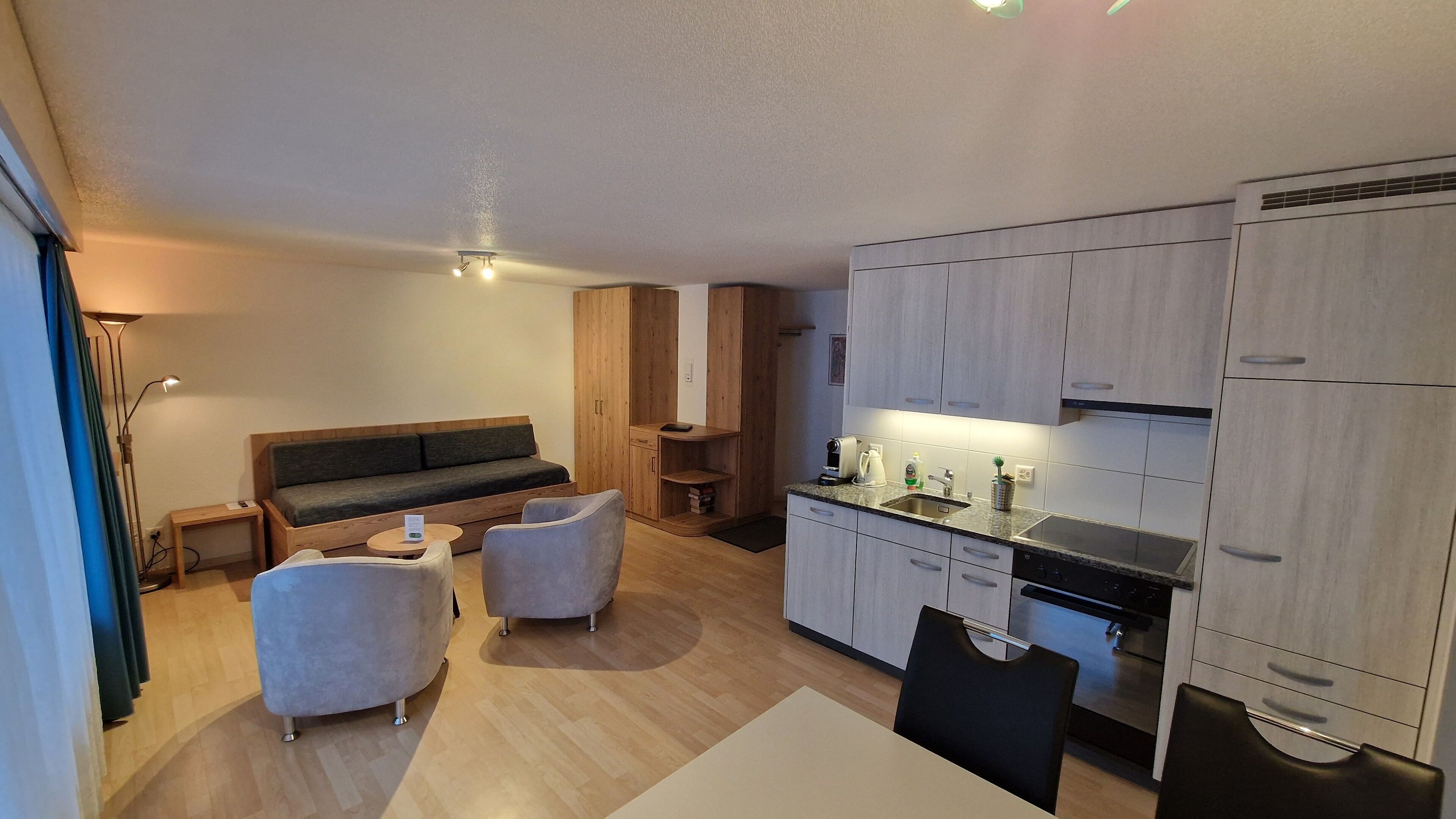 A modern, compact living space with a sofa, two armchairs, a small dining table, and a kitchenette.