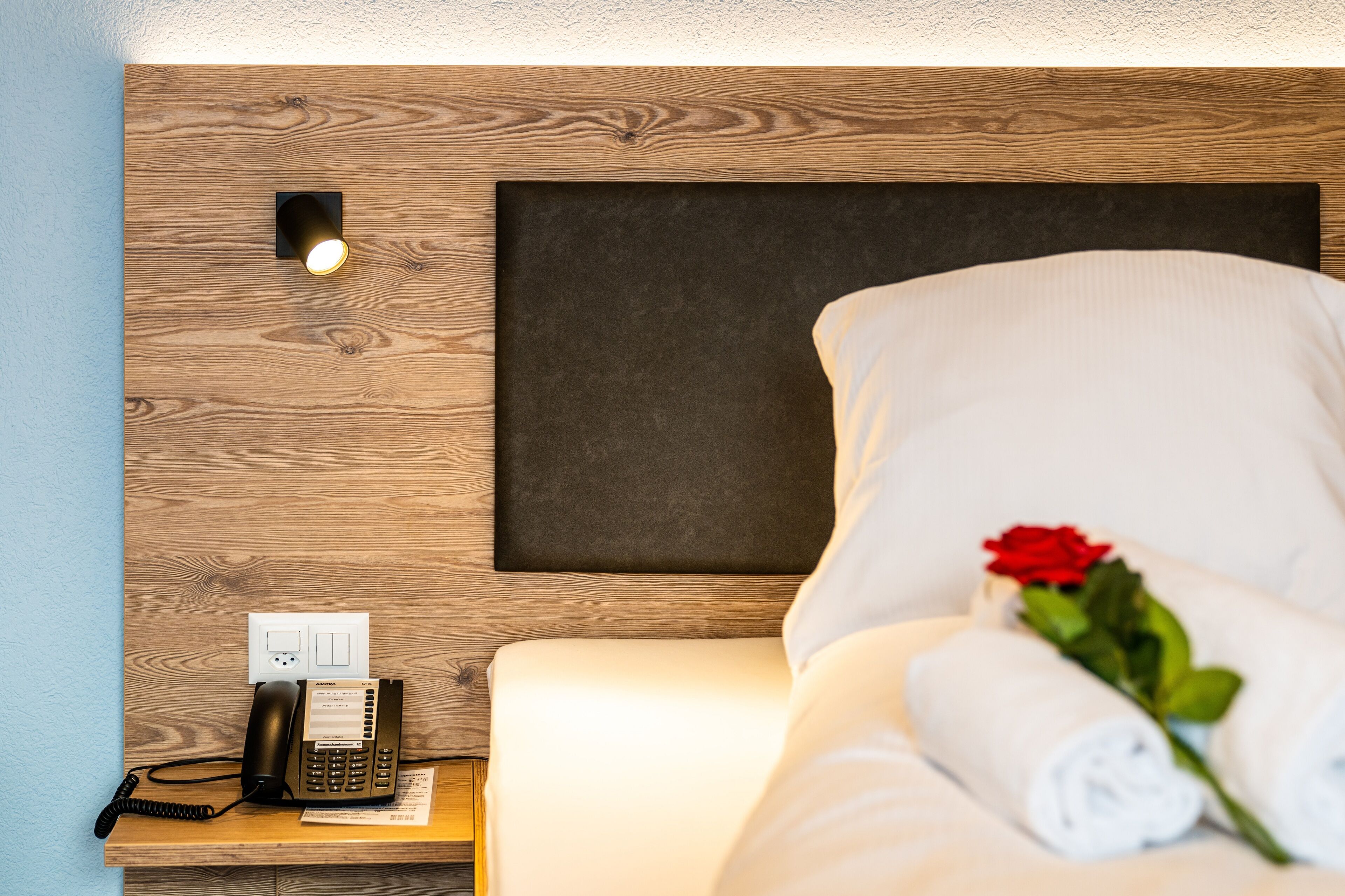 A hotel room with a wooden headboard, a bedside phone, a rolled towel, and a red rose.