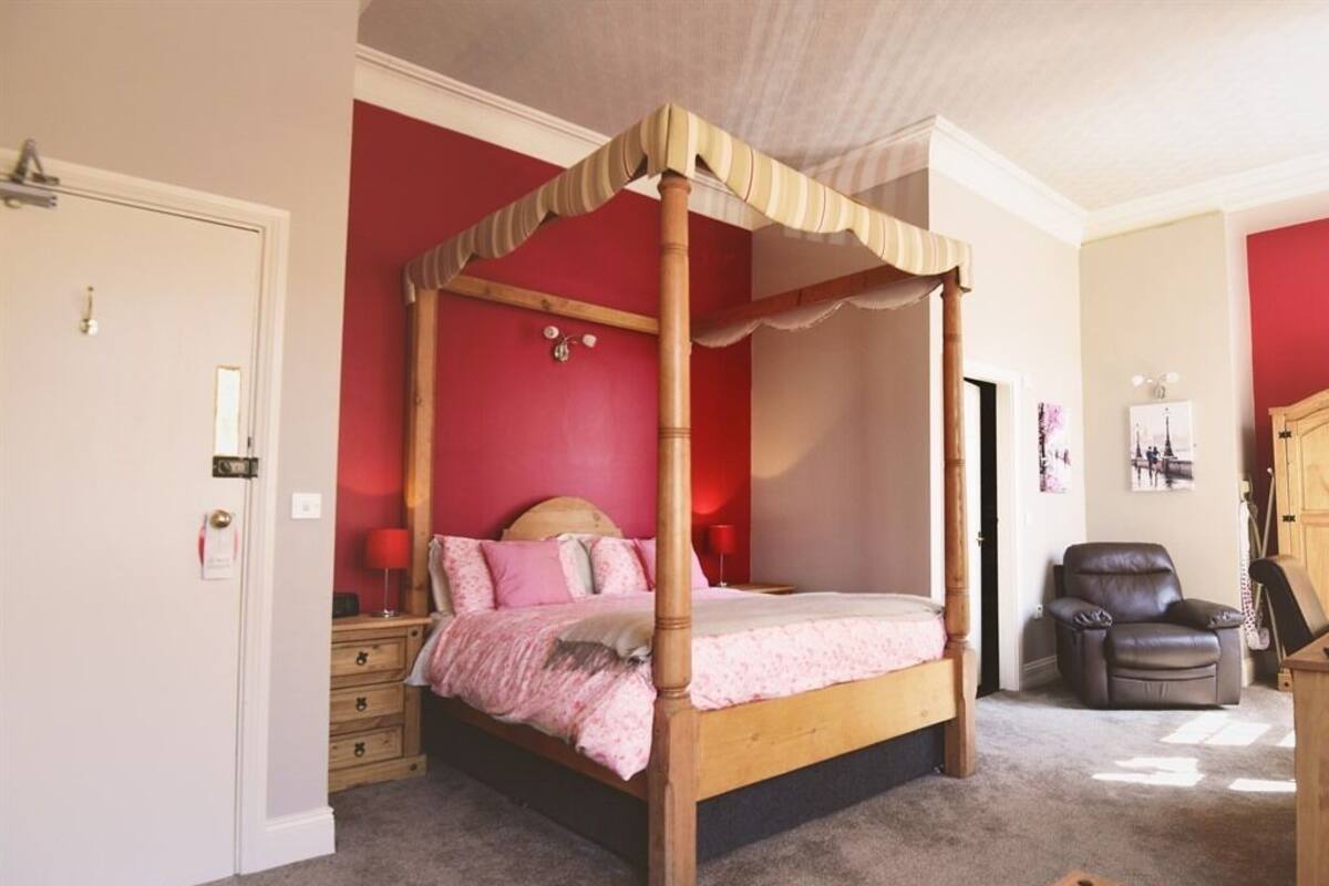 A four-poster bed with a canopy, a wooden nightstand, a chair, and a bedside table with a tissue box.