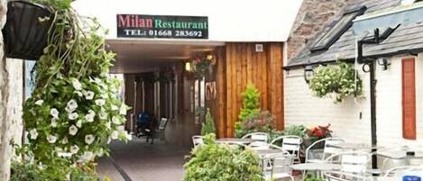 An outdoor seating area with tables and chairs, a sign for Milan Restaurant, and various potted plants.