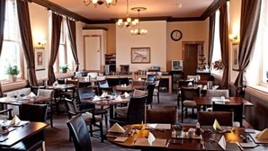 A hotel dining area with multiple tables and chairs, chandeliers, and a clock on the wall. - Black Bull Inn (Wooler)