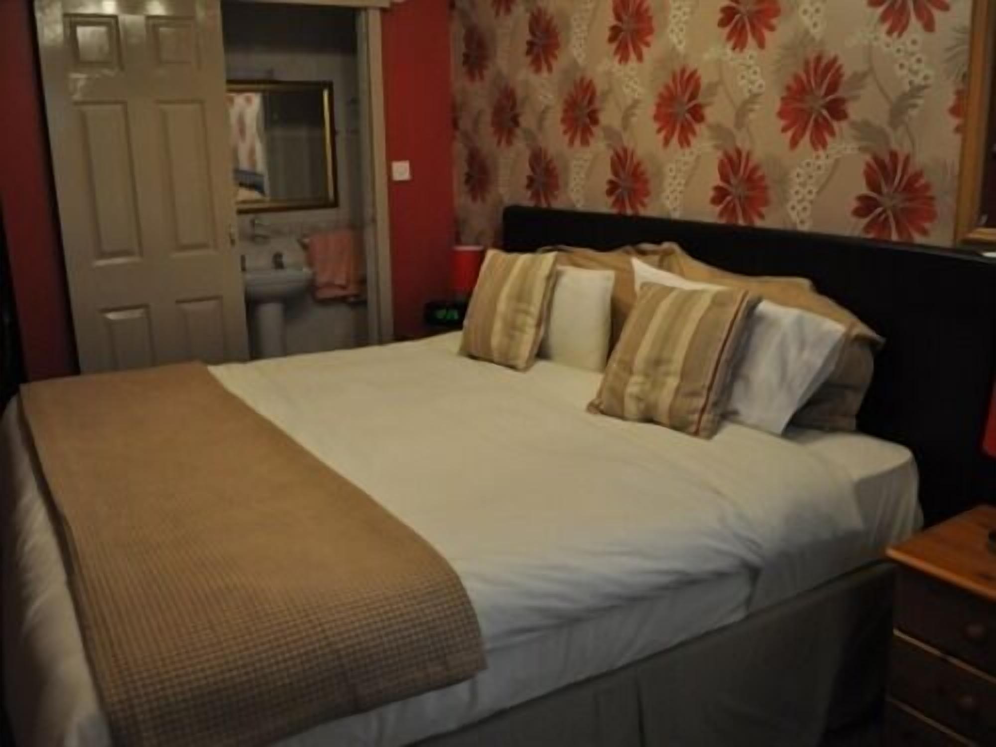 A bed with a beige blanket and pillows, a wooden nightstand, and a floral-patterned wall.