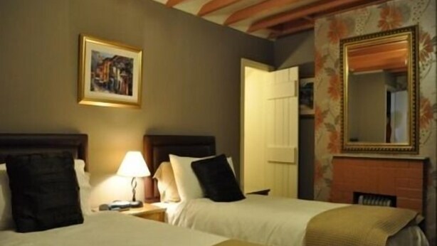 A hotel room with two beds, a nightstand with a lamp, a framed picture on the wall, and a mirror above a fireplace. - Black Bull Inn (Wooler)