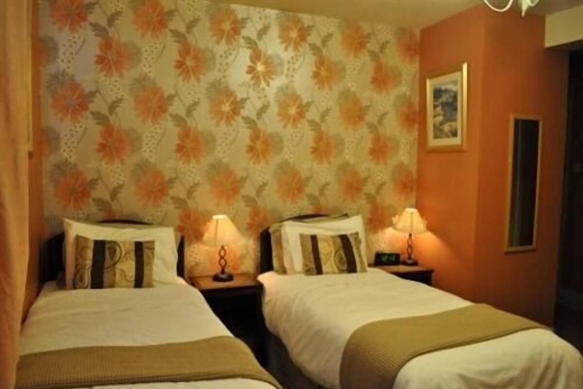 A room with two beds, each with a nightstand and a lamp. The walls have a floral wallpaper pattern.