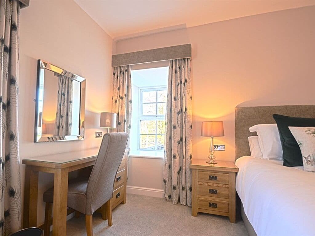 A hotel room with a bed, a desk, a chair, a nightstand with a lamp, and a window with curtains.
