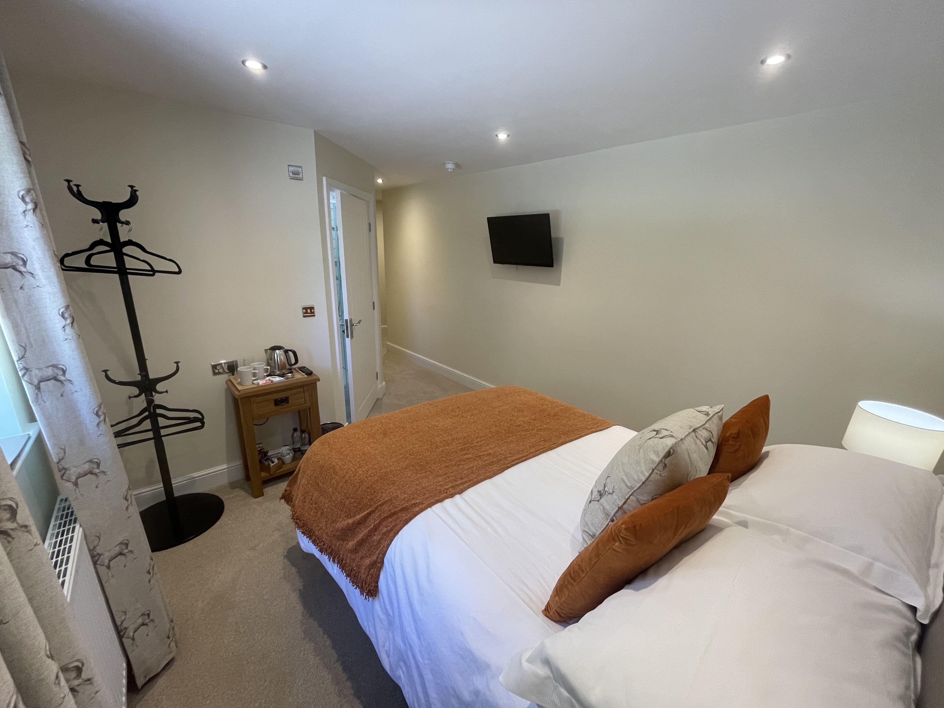Un dormitorio con una cama, una mesita de noche con lámpara, un perchero y un televisor en la pared.