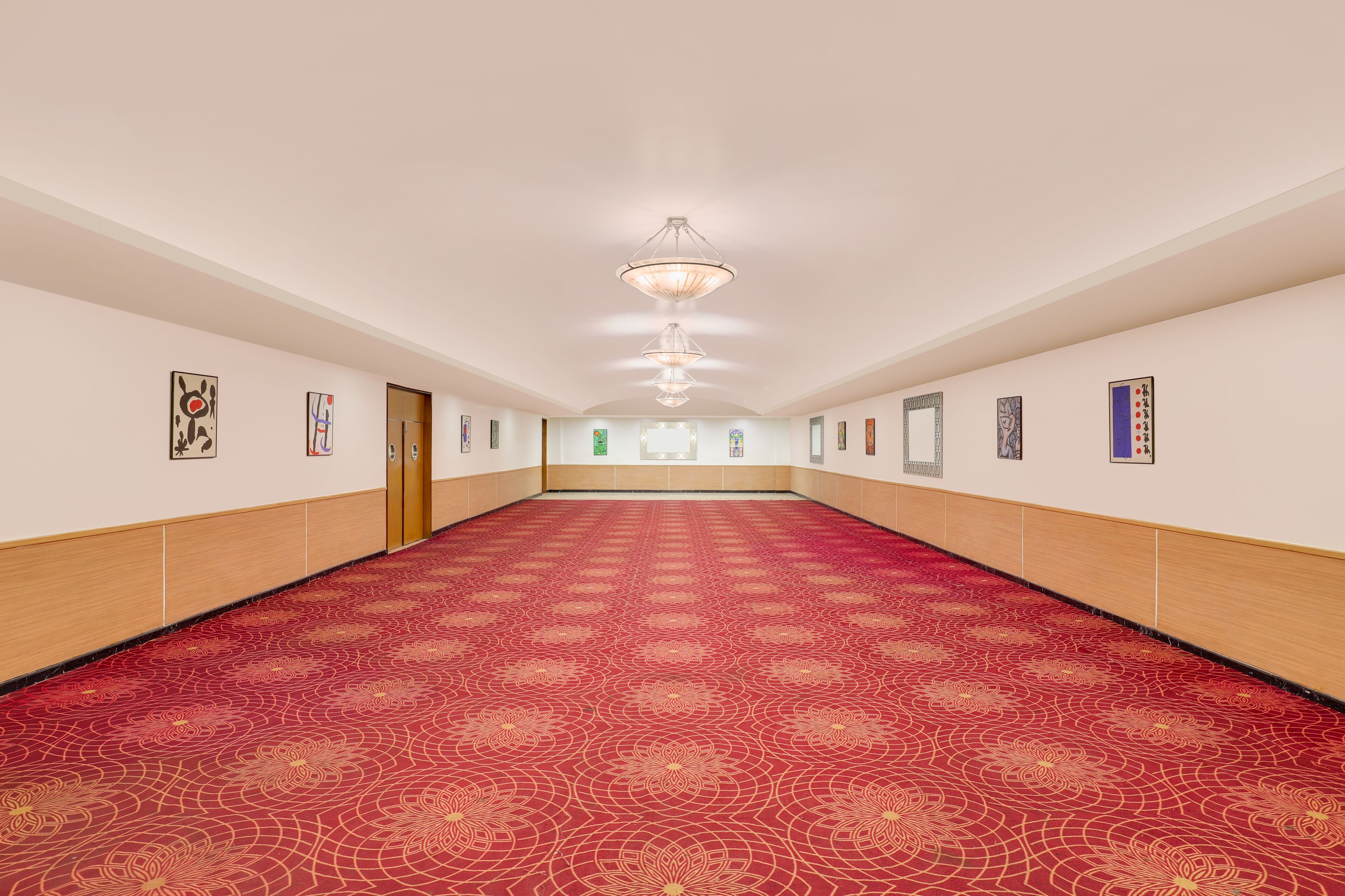 Habitación de hotel alargada y estrecha, con alfombra roja estampada, paneles de madera en las paredes y varias obras de arte enmarcadas.