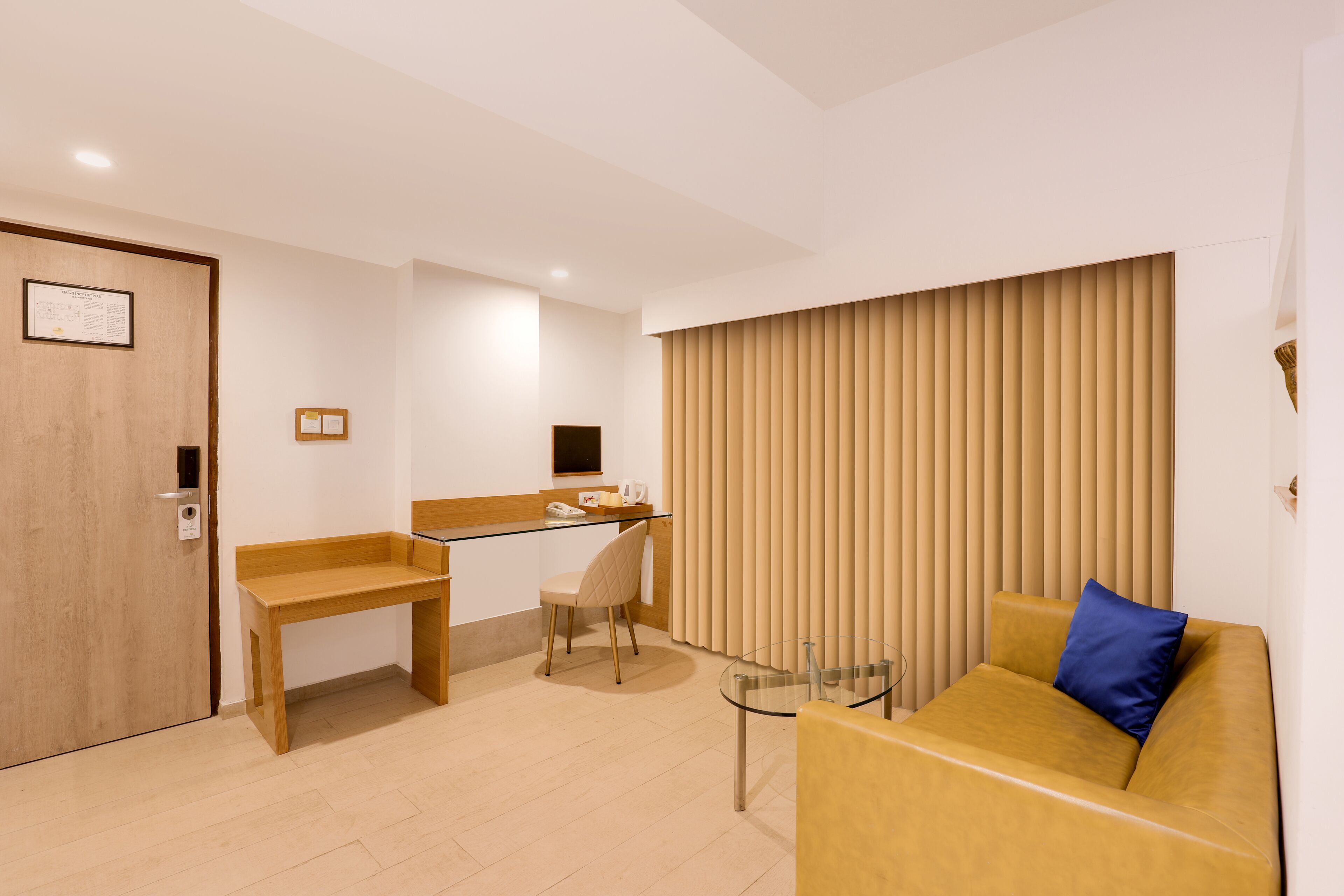 A modern hotel room with a wooden desk, a glass table, a yellow sofa with a blue pillow, and vertical blinds on the window.