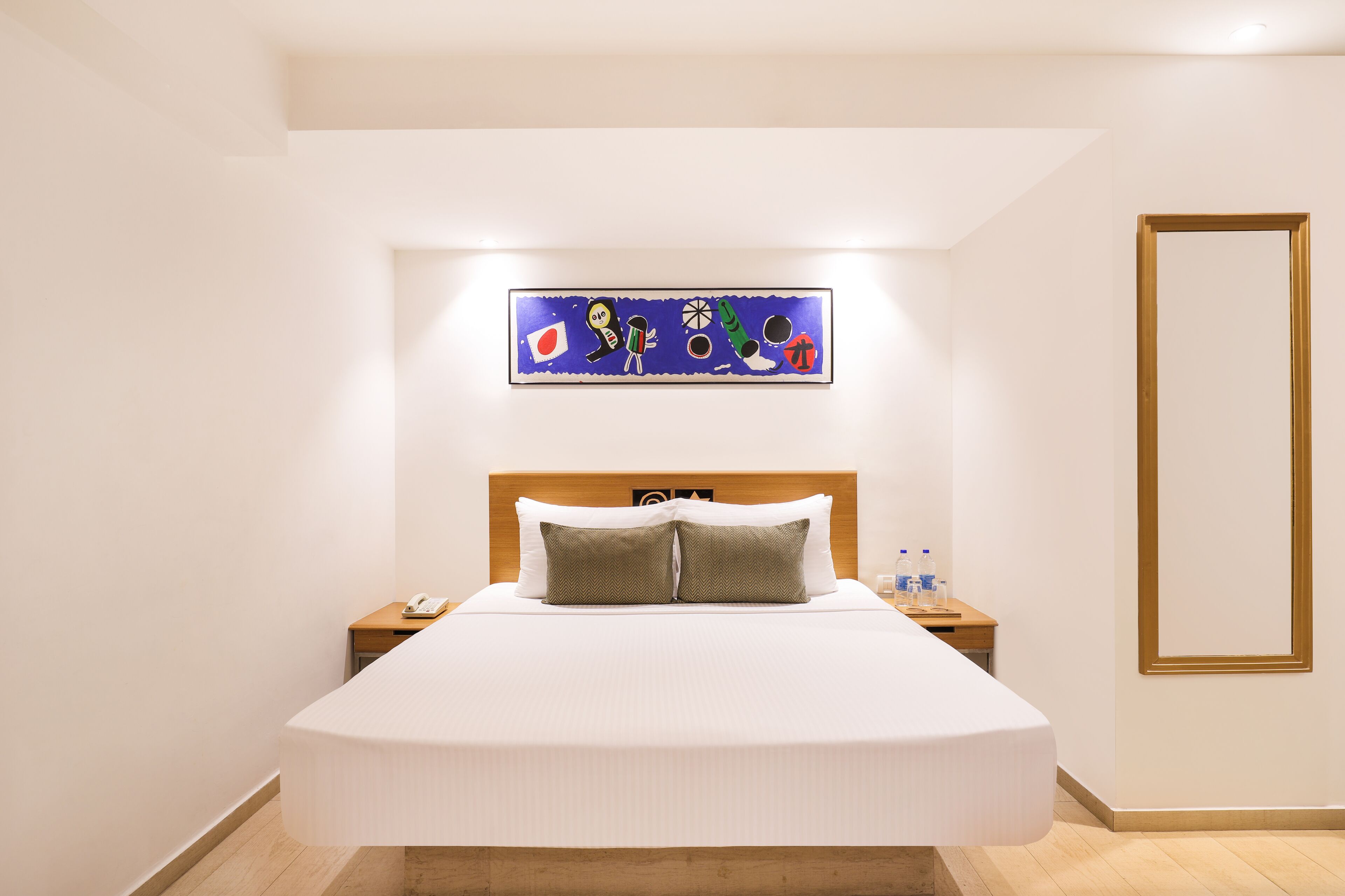 Una habitación de hotel con una cama grande, una mesita de noche con un teléfono, una mesita junto a la cama con botellas de agua y una obra de arte en la pared sobre la cama.