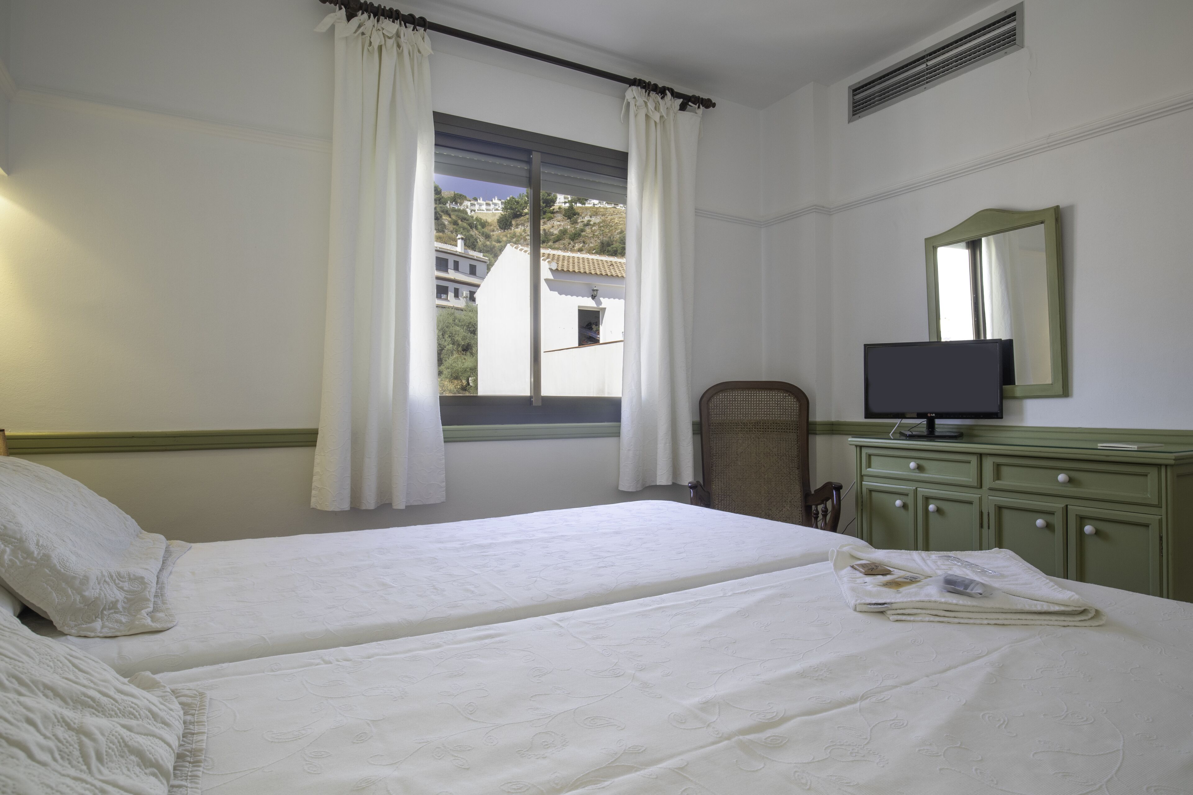 Une chambre avec deux lits, un téléviseur posé sur une commode et une fenêtre avec des rideaux.
