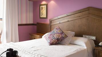Una cama bien hecha con edredón blanco y un cojín decorativo, cabecera de madera, mesita de noche con lámpara y pared con estampado a rayas.