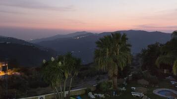 A swimming pool with a circular design, surrounded by lounge chairs and palm trees, set against a backdrop of mountains and a twilight sky.