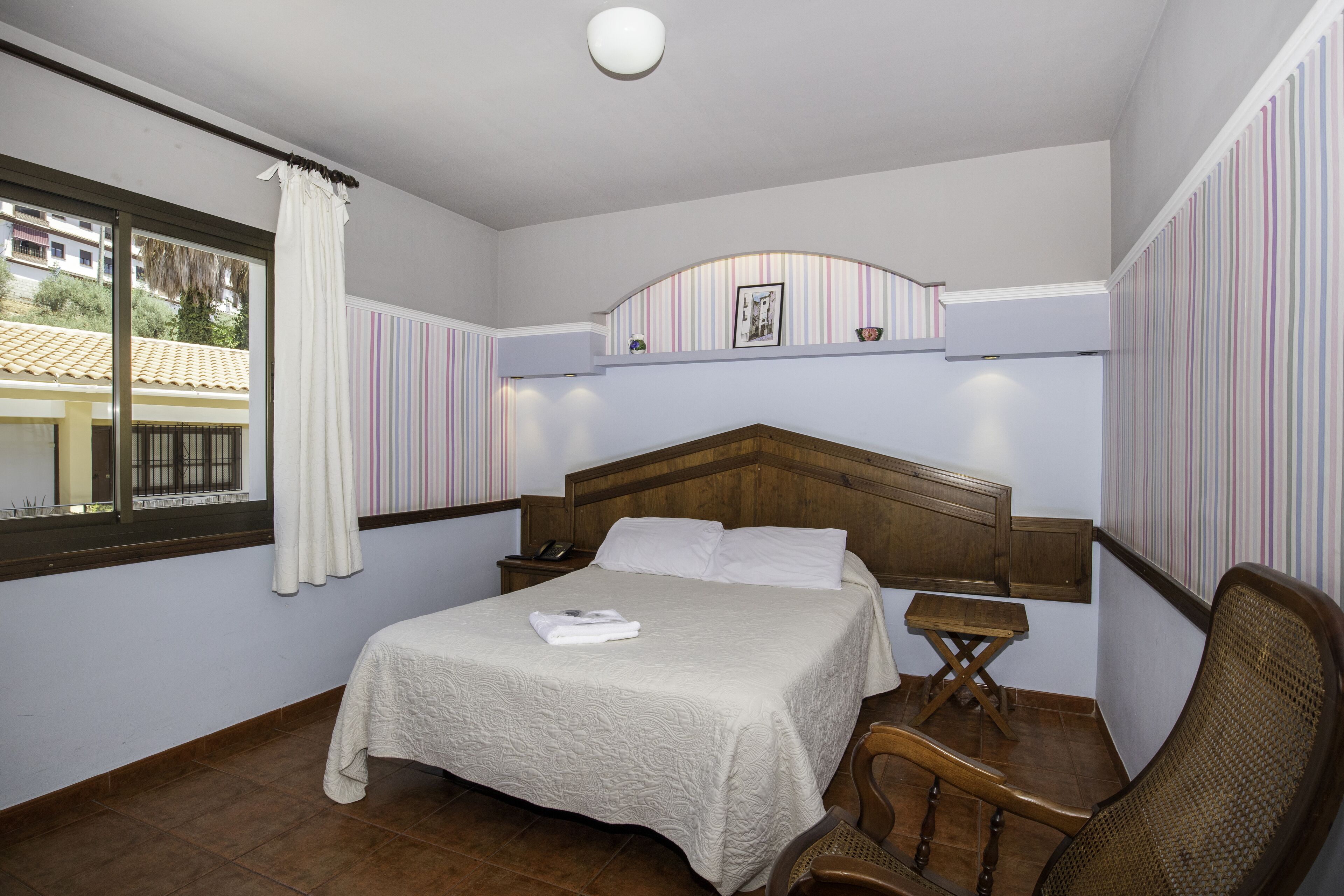 A bedroom with a bed, a bedside table, a chair, a window with curtains, and striped wallpaper.