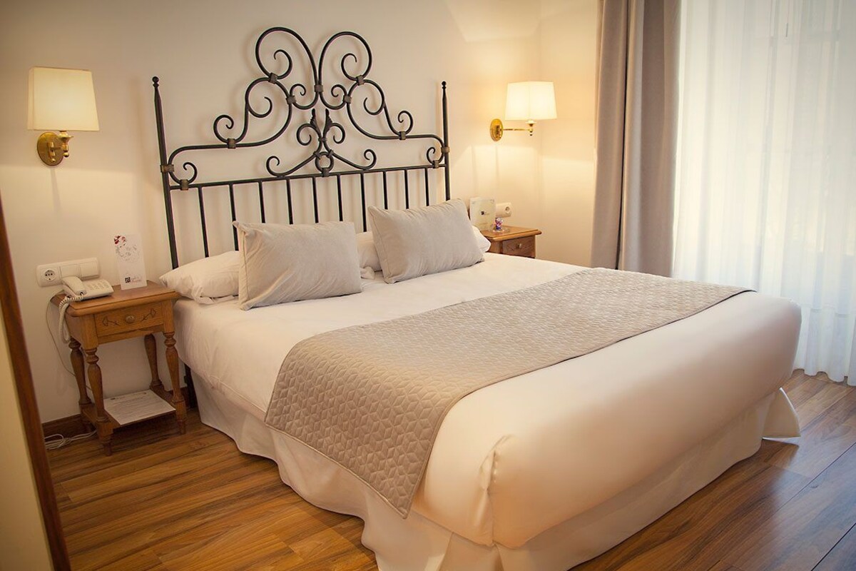 Una cama bien tendida con un cabecero decorativo, dos mesitas de noche con lámparas y suelo de madera.
