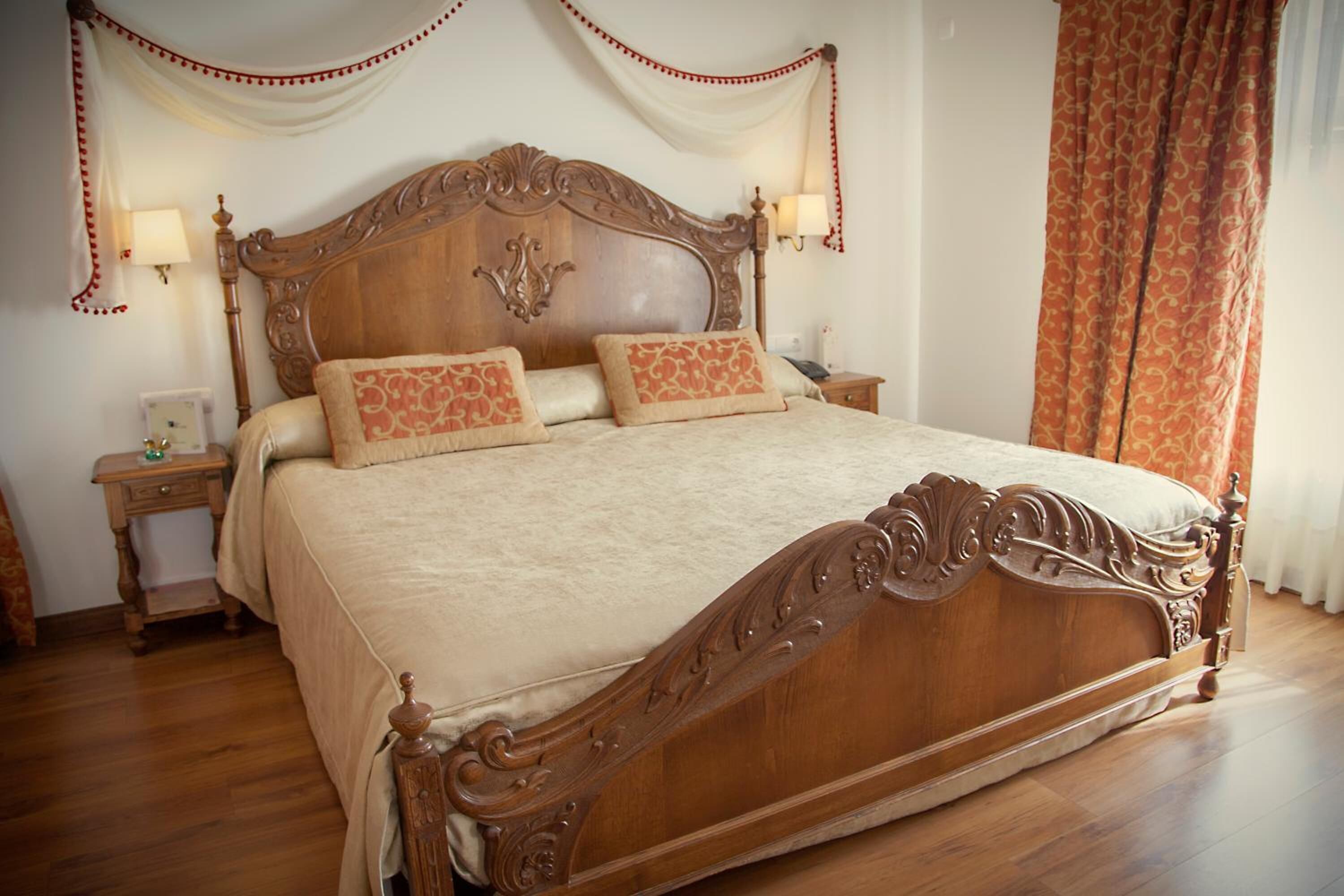 Una cama de madera grande con adornos labrados, dos mesitas de noche con lámparas y una cortina en la ventana.
