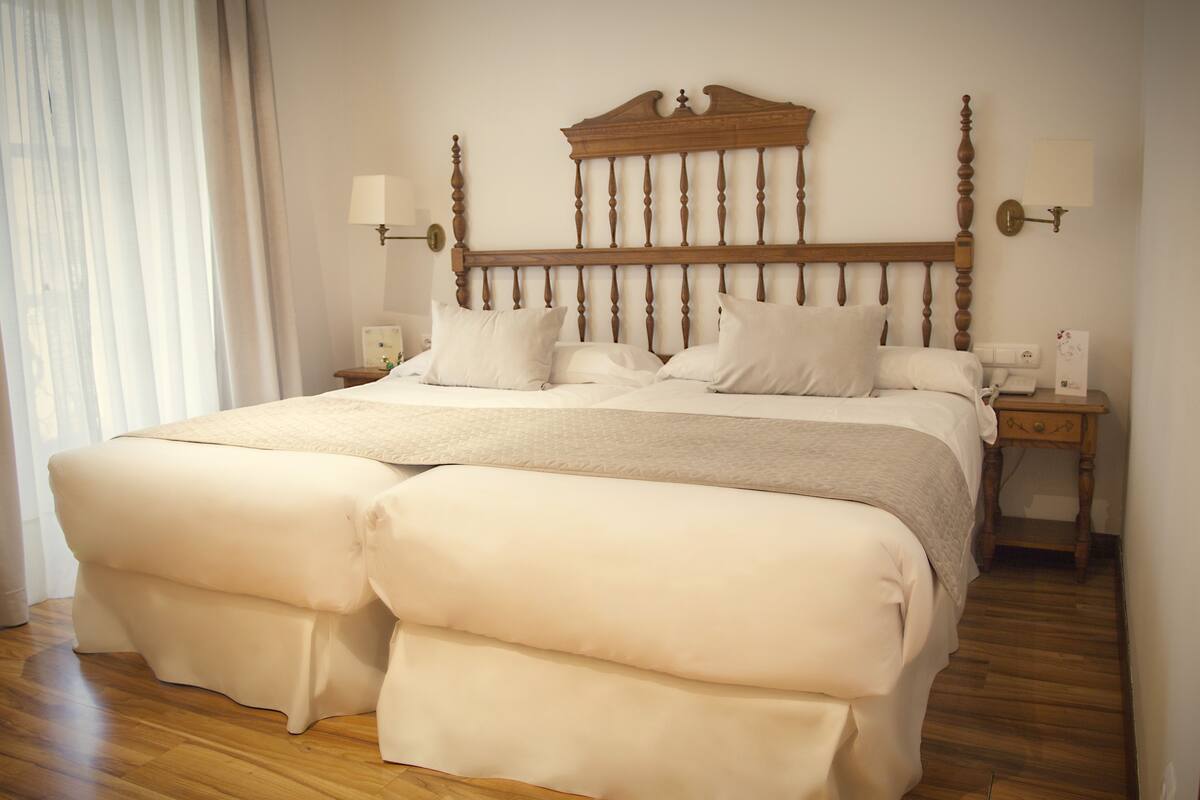 A neatly made bed with a wooden headboard, a bedside table with a lamp, and a nightstand with a phone and a card.