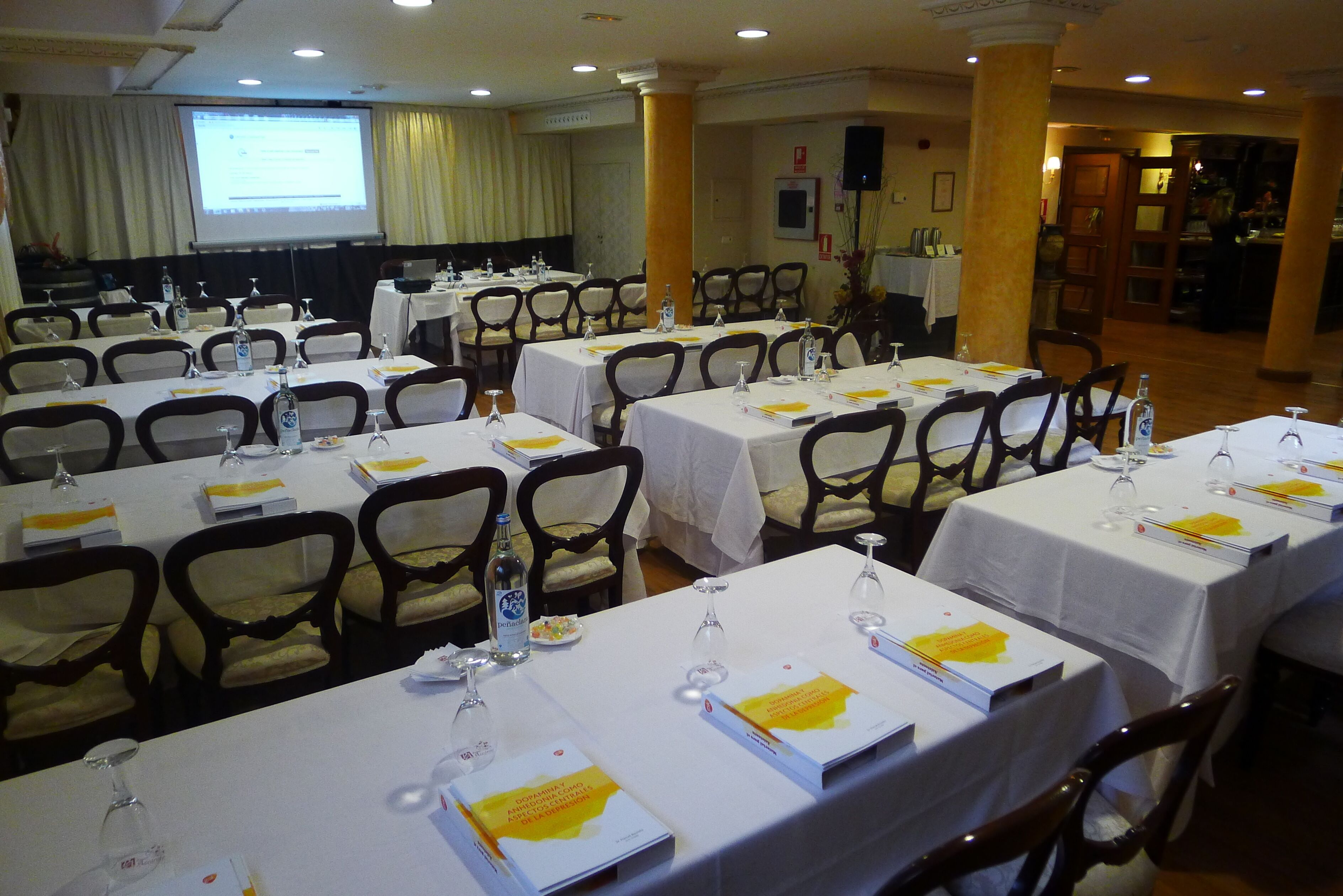 Un salón de conferencias con varias mesas dispuestas para una reunión, cada una con placas de identificación, botellas de agua y documentos.