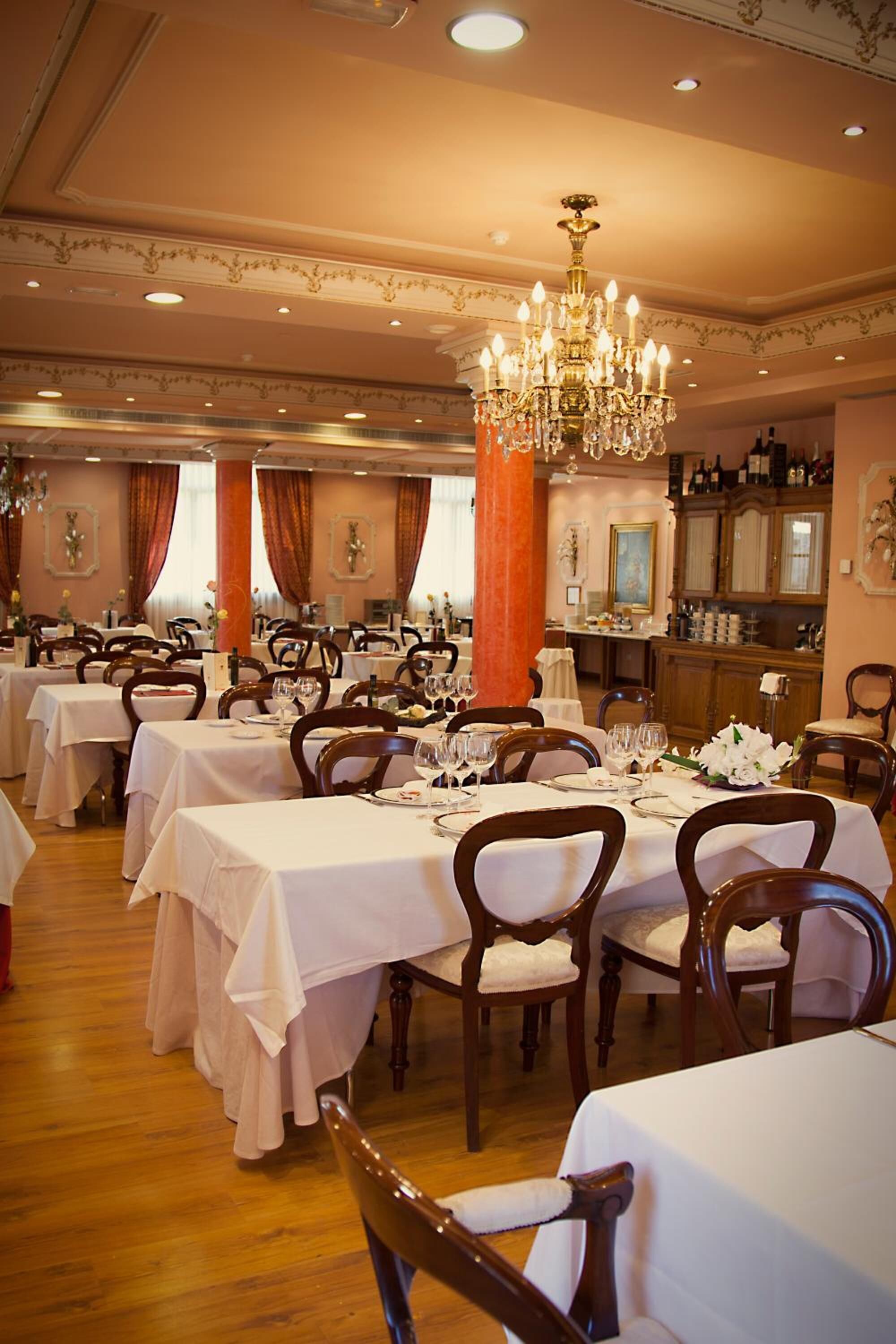 A formal dining room with multiple tables set for service, a large chandelier, and a bar area in the background.