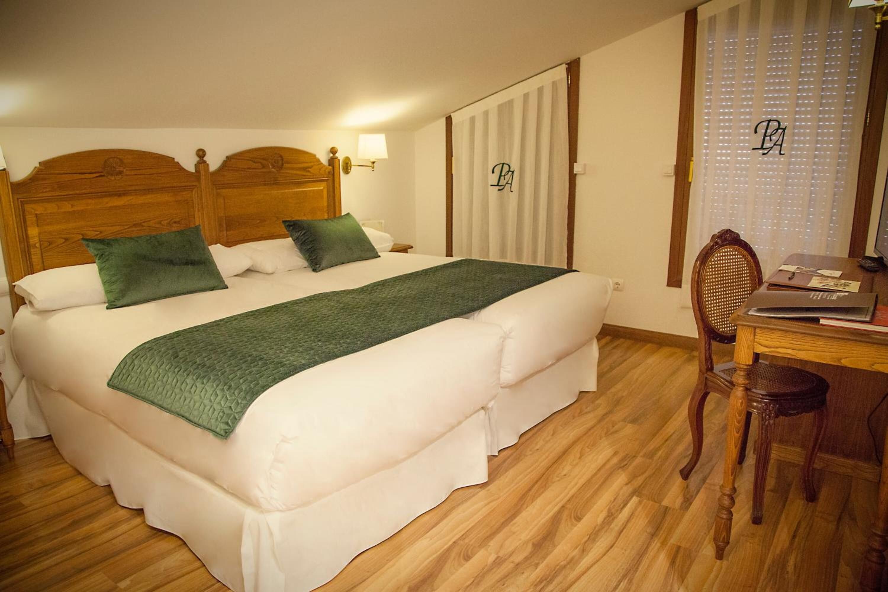 A neatly made bed with a wooden headboard, a green throw blanket, and two pillows. There is a wooden desk with a chair and a lamp.