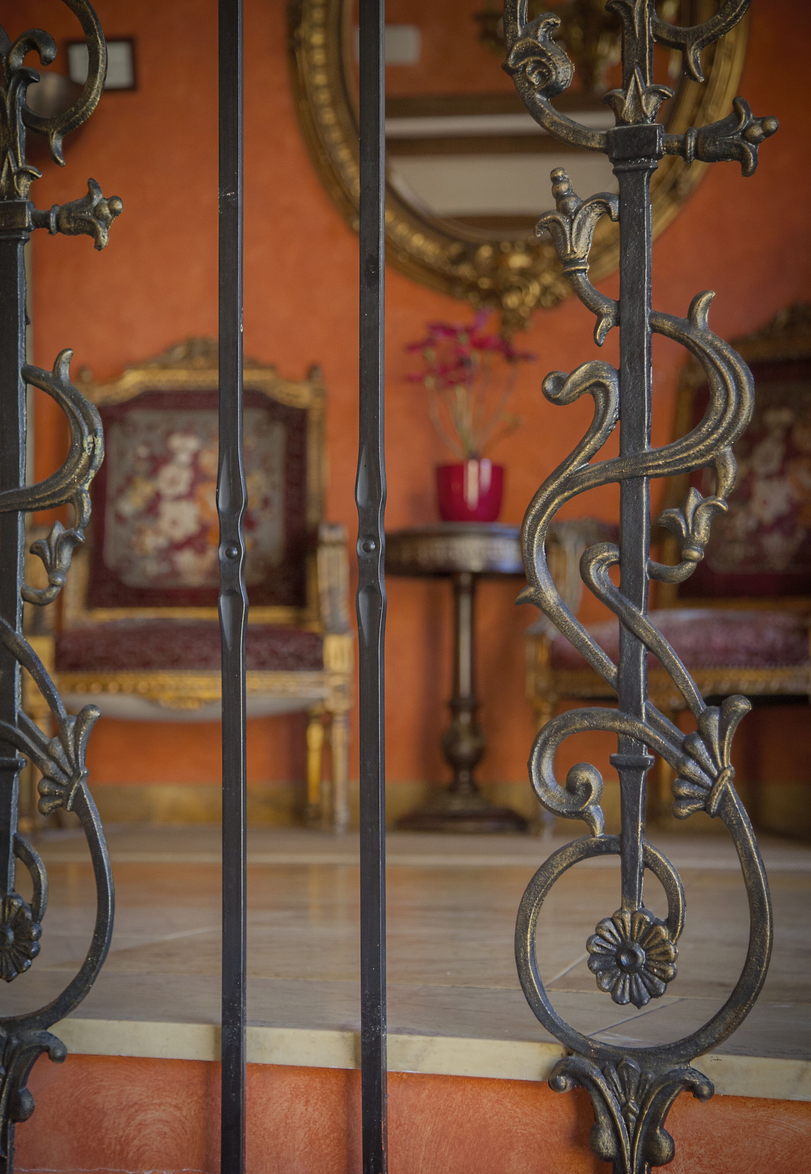 Des rampes de métal ouvragées aux motifs complexes, un vase rouge rempli de fleurs et un tableau encadré d’or accroché au mur.