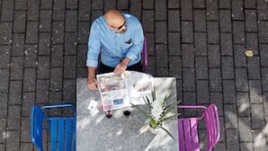 Un hombre sentado a una mesa con un periódico, rodeado de dos sillas.