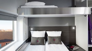 Una habitación de hotel moderna con literas, una mesita de noche con un libro y una ventana con vista a la ciudad.