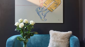 Een smaragdgroene bank met een zacht kussen, een vaas met witte bloemen en een boek op een klein zwart tafeltje. Achter op de muur hangt een grote ingelijste illustratie.