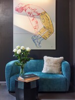 Een smaragdgroene bank met een zacht kussen, een vaas met witte bloemen en een boek op een klein zwart tafeltje. Achter op de muur hangt een grote ingelijste illustratie.
