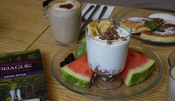 Uma mesa com um copo de iogurte coberto com granola e fruta, uma fatia de melancia, um livro intitulado "Praha Artel Style" e um prato com panquecas.