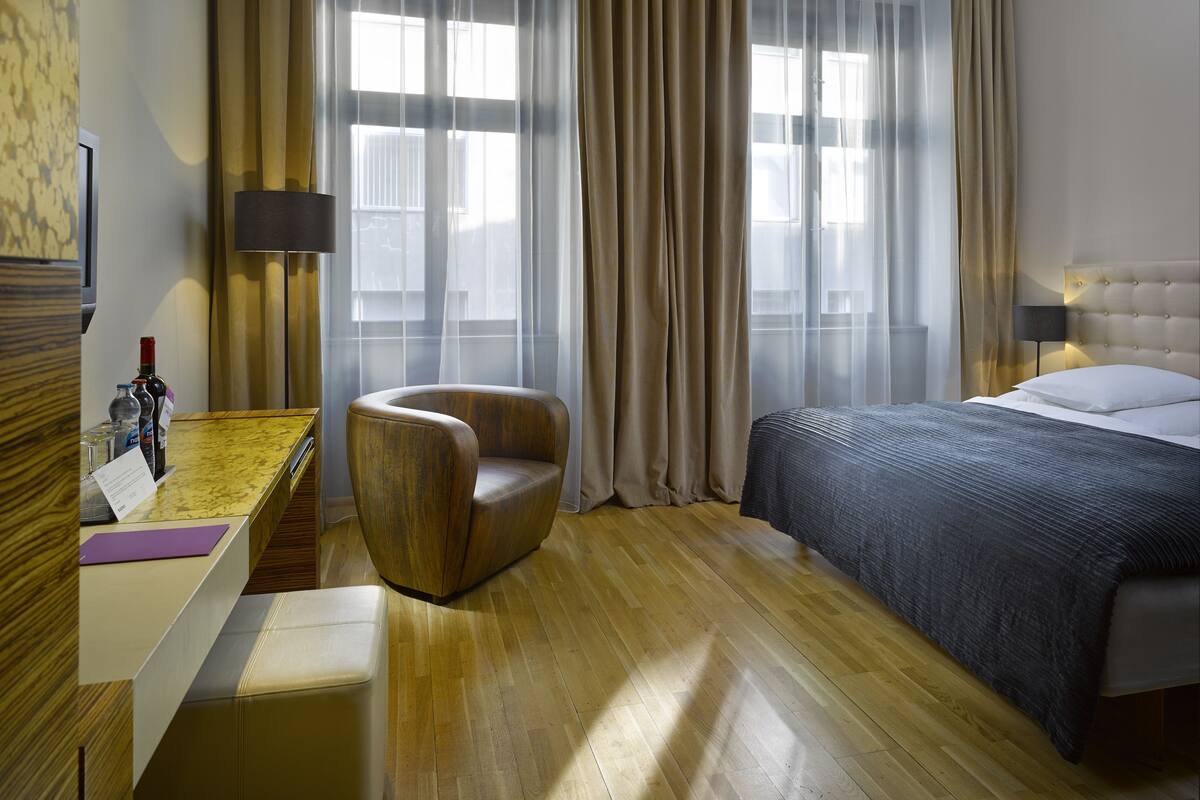 A hotel room with a bed, a chair, a desk with a lamp, and a view of the city through large windows.