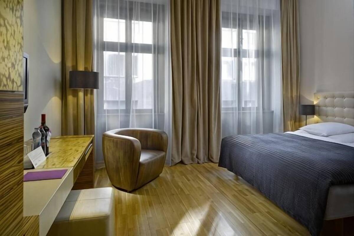 A hotel room with a bed, a chair, a desk, and a window with curtains.