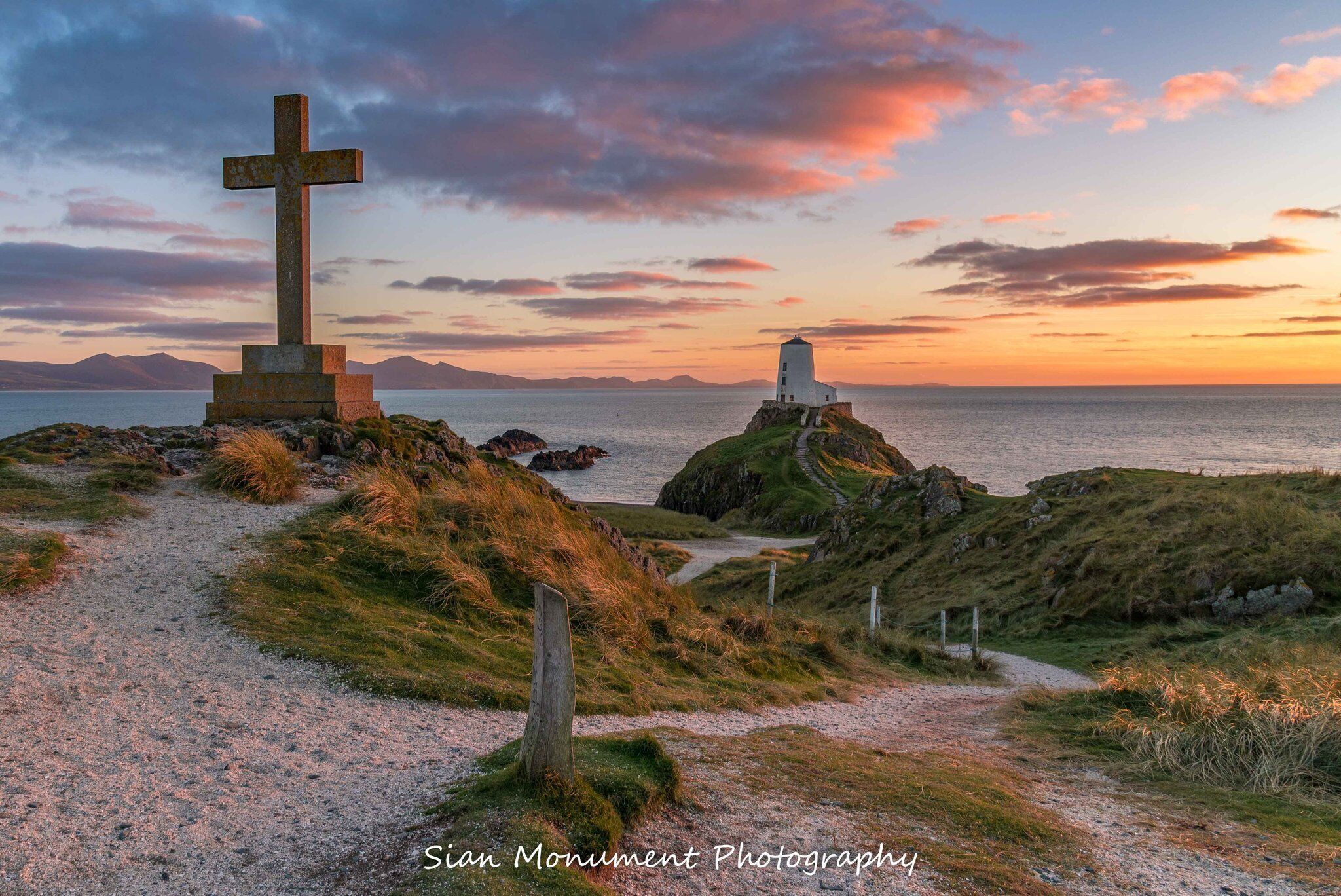 A cross monument and a lighthouse on a coastal path with a view of the sea and mountains in the background.